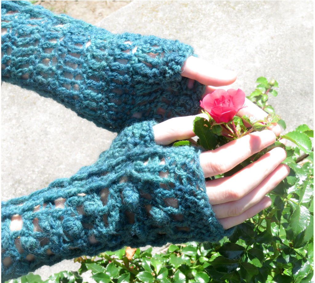 Close hands wearing crochet lace fingerless mitten worked in blue.  Hands gently hold rosebud